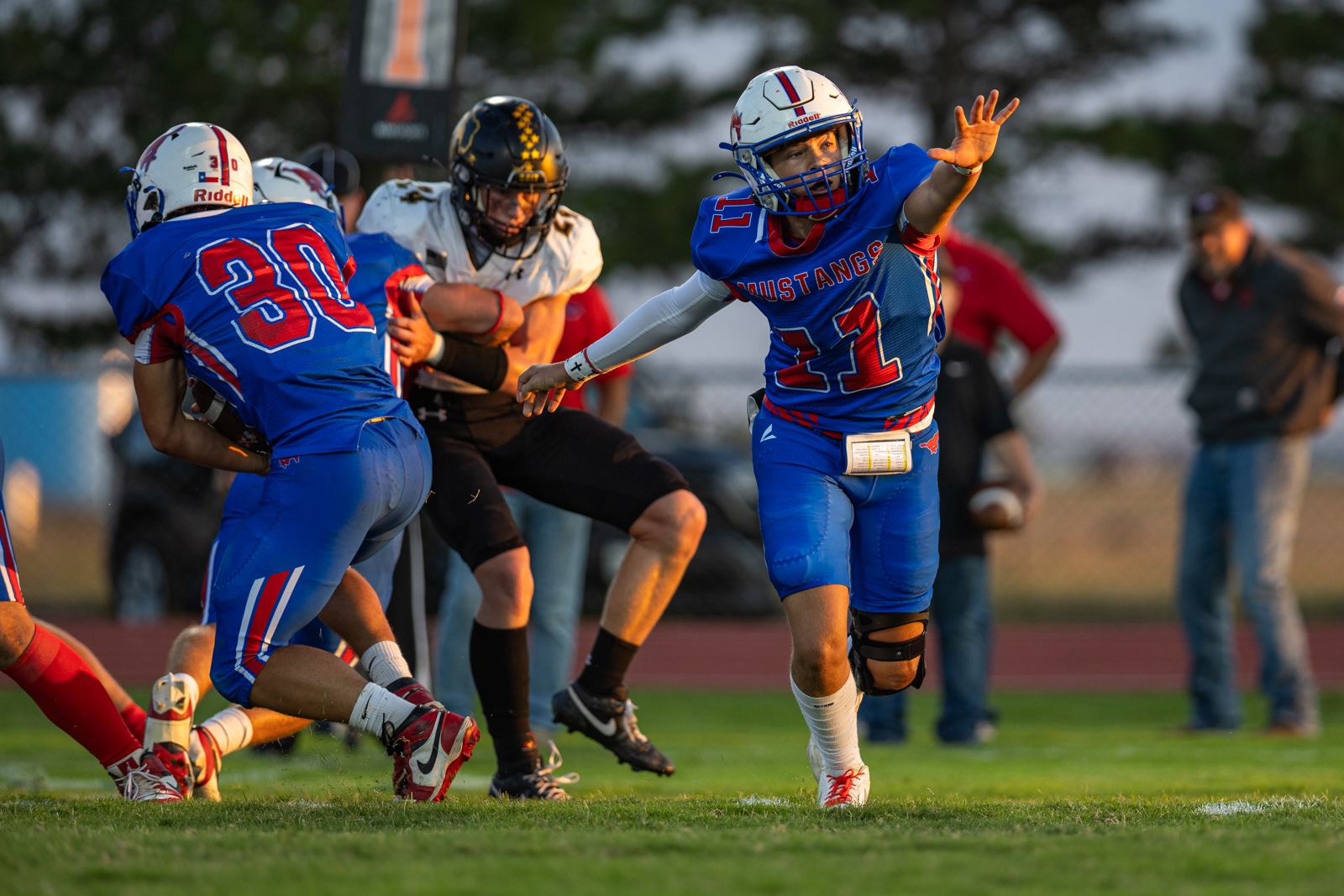 Liberty_15-22 | Press Pass Sports American football action: blue‑uniform Mustang player (#11) extends an arm as teammates block nearby and an opposing player engages in the scrum on the grass field.