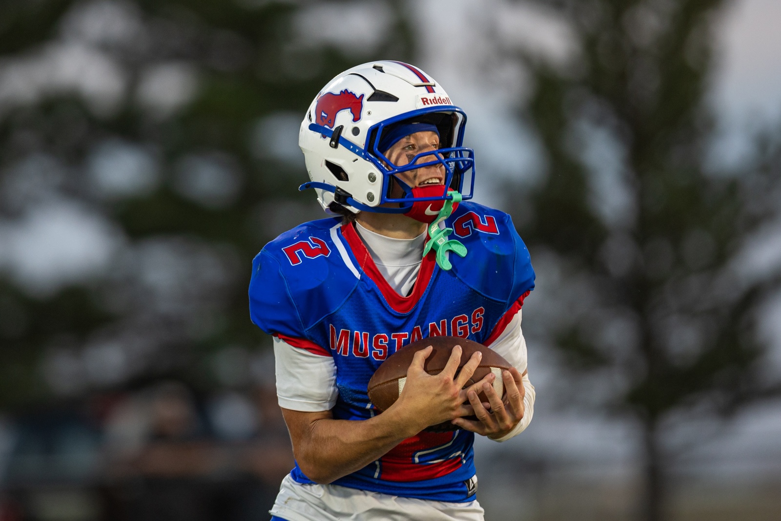 Liberty_15-24 | Press Pass Sports American football player in a blue Mustangs jersey catches or holds a football, wearing a white helmet with a red logo and blue face mask; motion on-field.