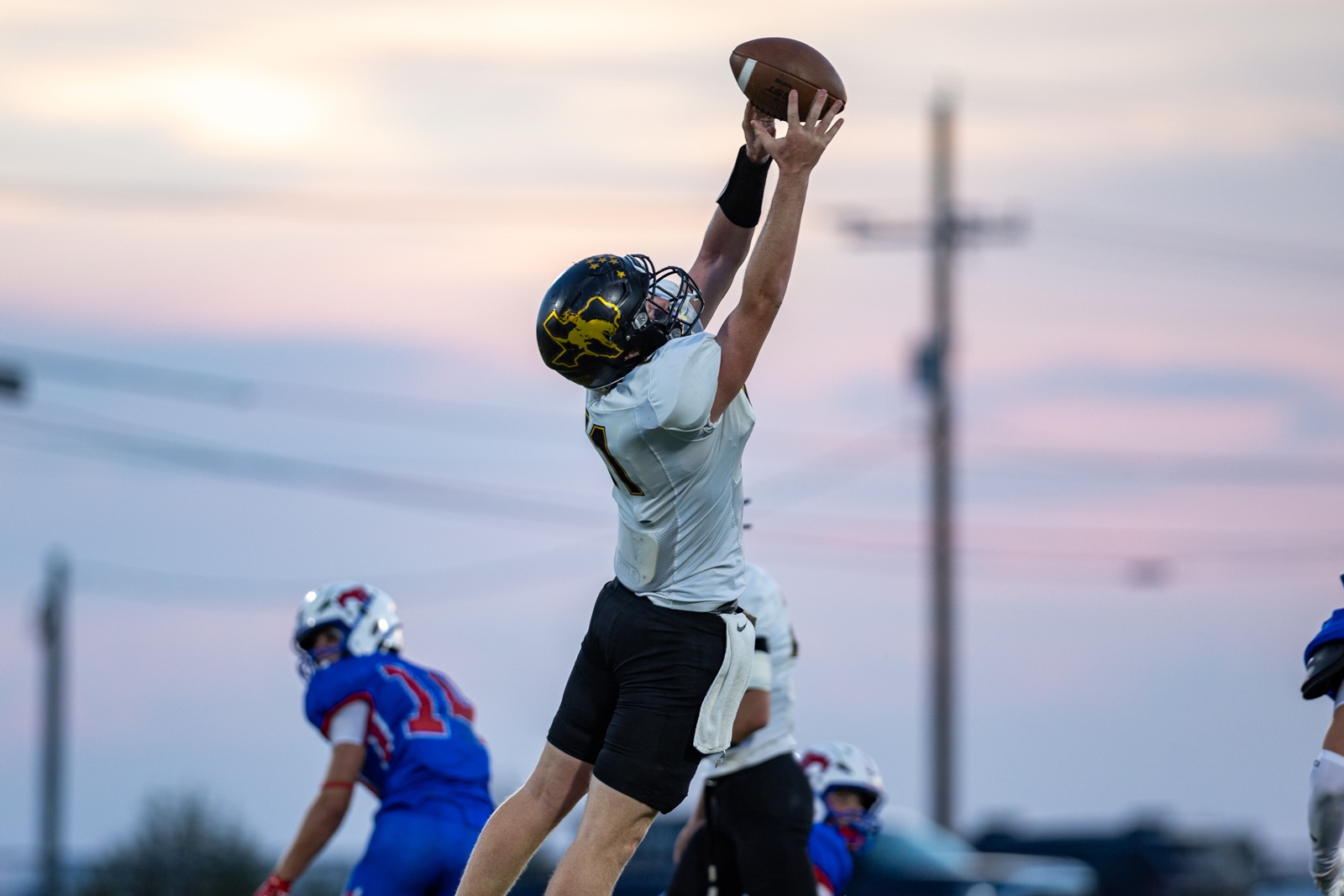 Liberty_15-26 | Press Pass Sports Football player in a white jersey jumps to catch a pass during a game at sunset/orange-pink sky behind.