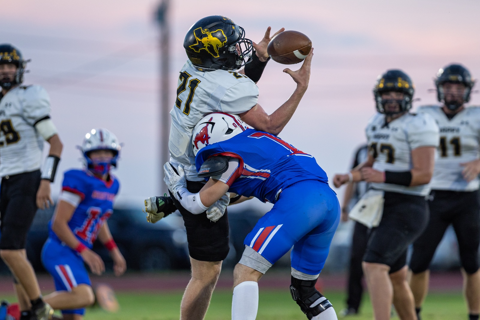 Liberty_15-27 | Press Pass Sports Football action: a white-jersey player leaps to catch the ball while a blue-jersey defender collides into him from below on a sunny field.