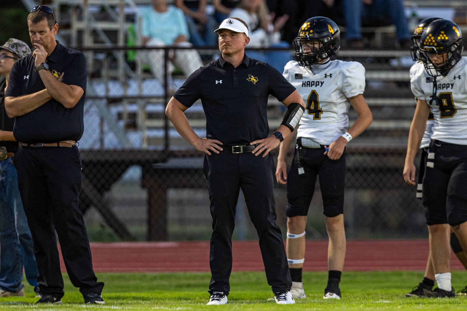 Liberty_15-29 | Press Pass Sports Coach in black polo with white visor stands on the sideline with hands on hips while players in white uniforms watch nearby on the field.