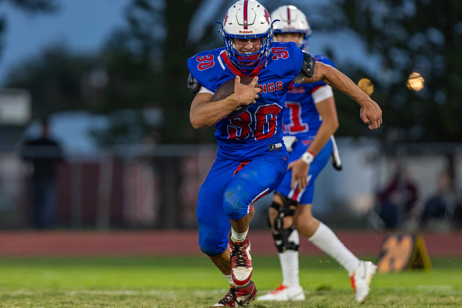 Liberty_15-30 | Press Pass Sports American football player in blue uniform sprinting with ball tucked under his arm on a grassy field during a game