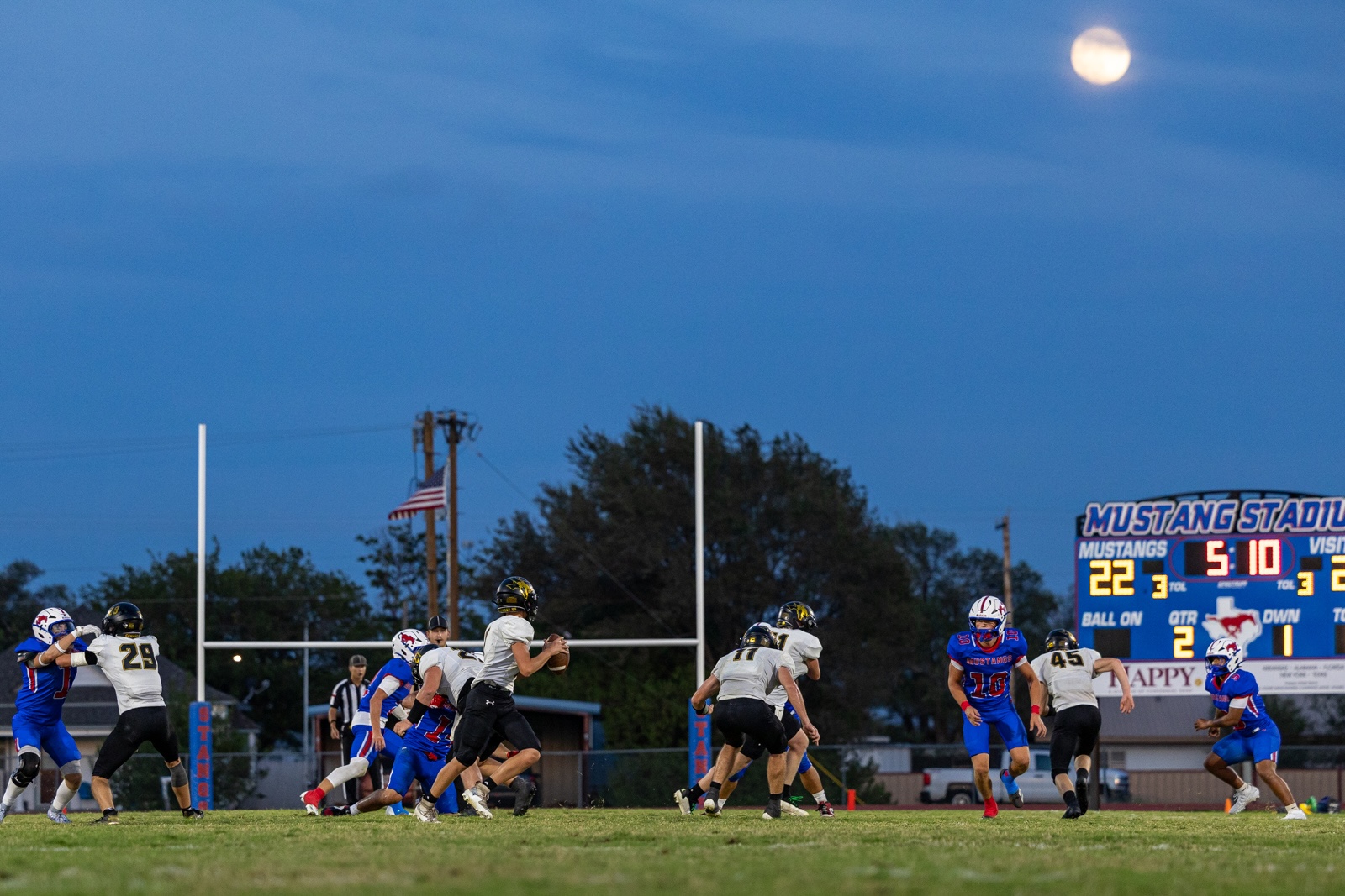 Liberty_15-32 | Press Pass Sports Football game on a bright field at dusk with a full moon overhead; players in white, black, and blue uniforms collide near the goal posts, Mustang Stadium scoreboard in the background.
