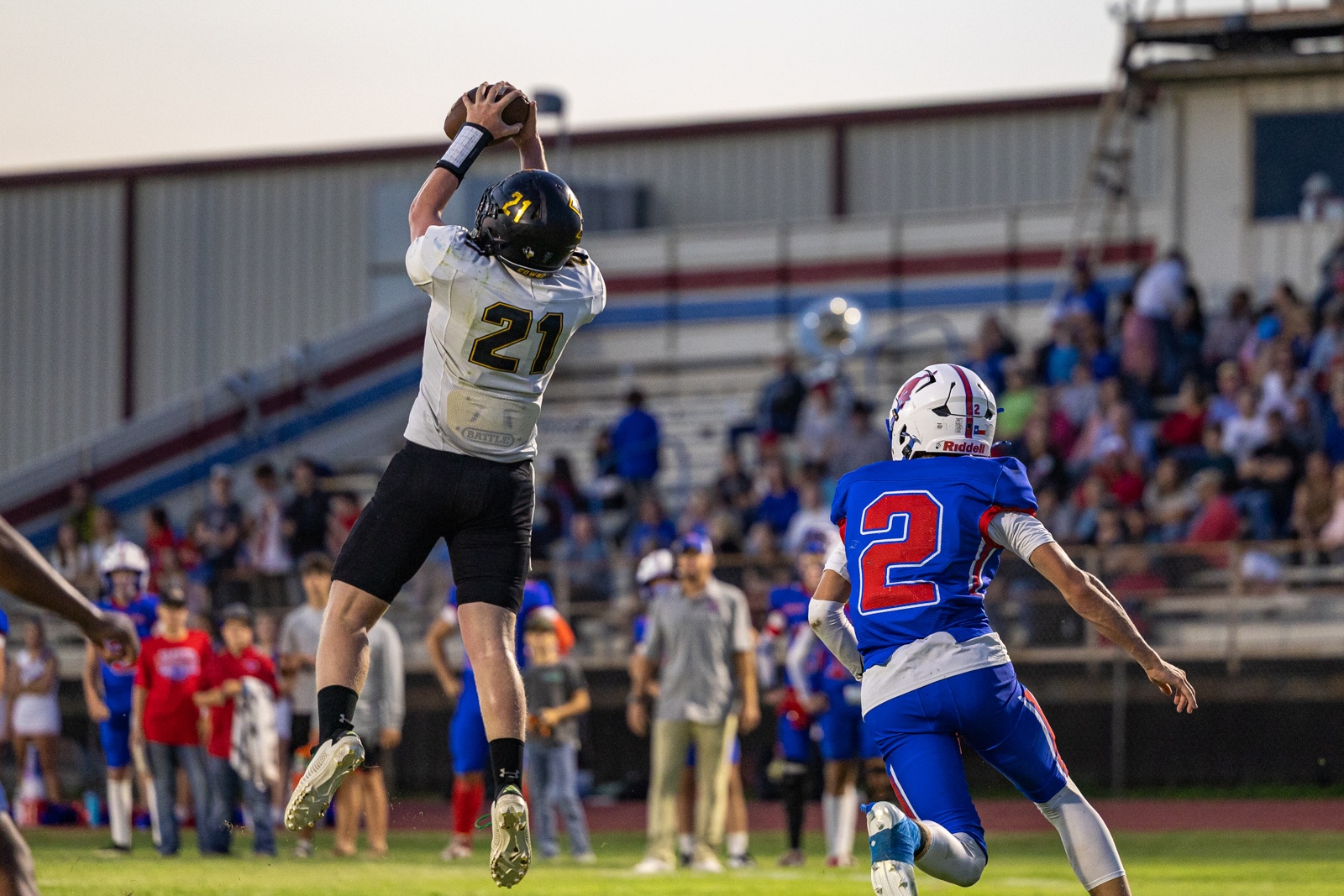 Liberty_15-33 | Press Pass Sports Football player in white jersey #21 leaps to catch the ball over a defender in blue #2 on a sunny stadium field.