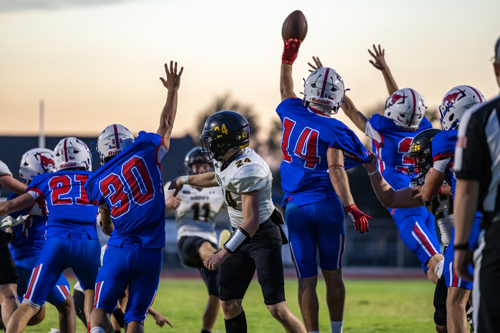 Liberty_15-34 | Press Pass Sports Football game with players in blue and white and a central white-uniformed player reaching for the ball amid a high-tive scrum on the field at sunset variants of play