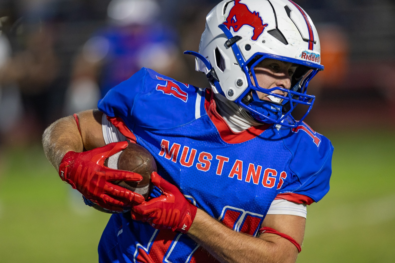 Liberty_15-36 | Press Pass Sports Football player in a blue Mustang uniform clutching a football, wearing a white helmet with a red mustang logo, mid-run on a field.