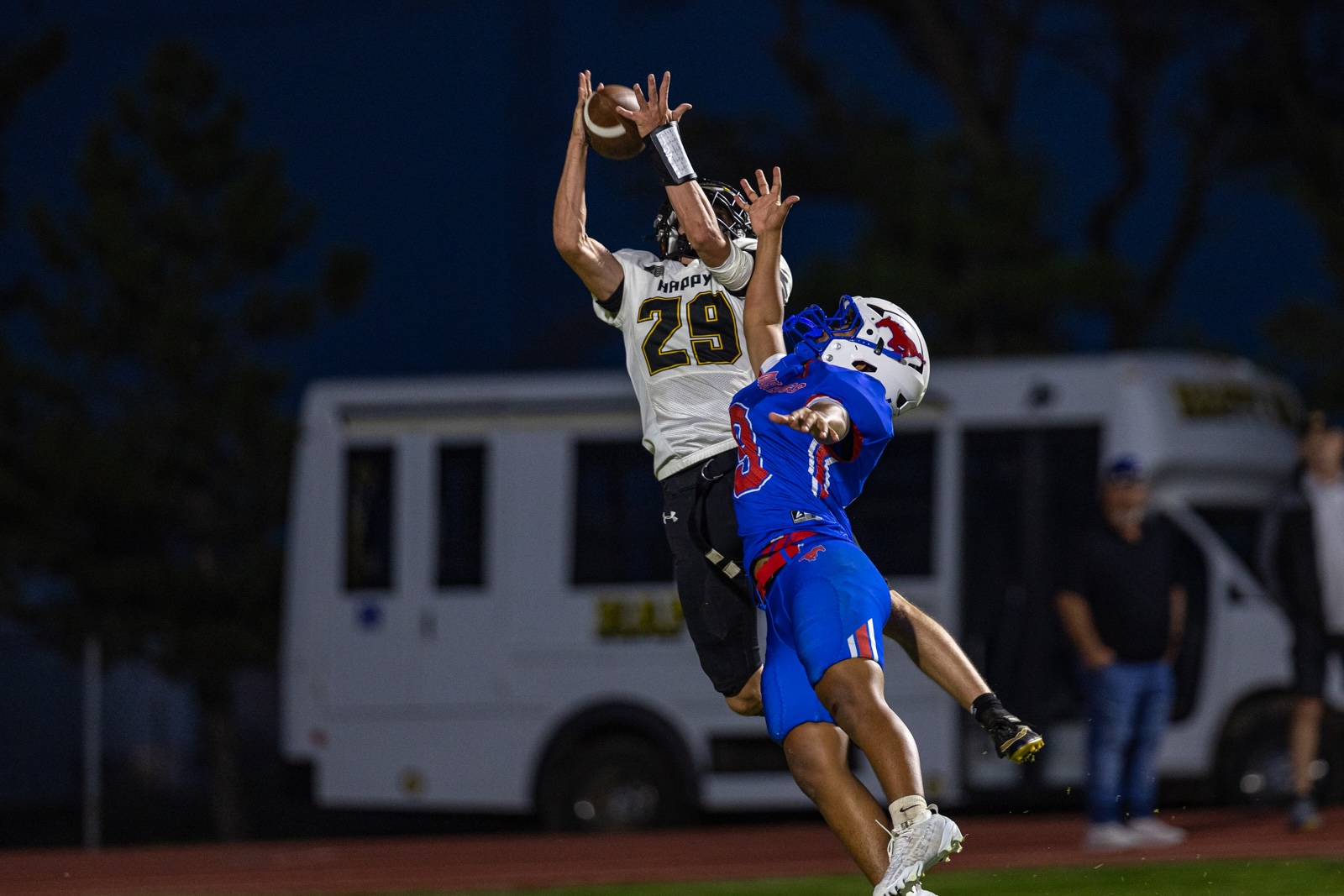Liberty_15-37 | Press Pass Sports Two football players jump to contest a catch at night; one in a white jersey number 29 reaching for the ball while a blue-clad defender reaches up