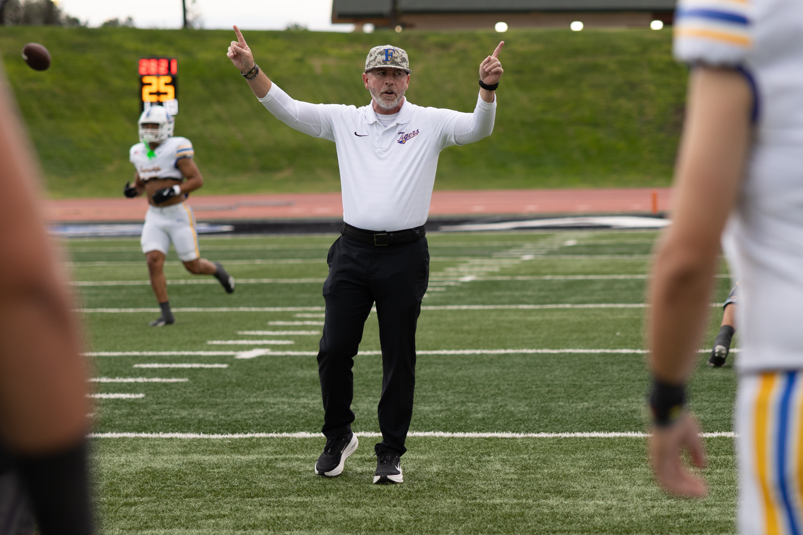 007A5598 | Press Pass Sports Football coach wearing a white polo and camo hat signals a play, arms raised and pointing on a football field with players in white uniforms nearby