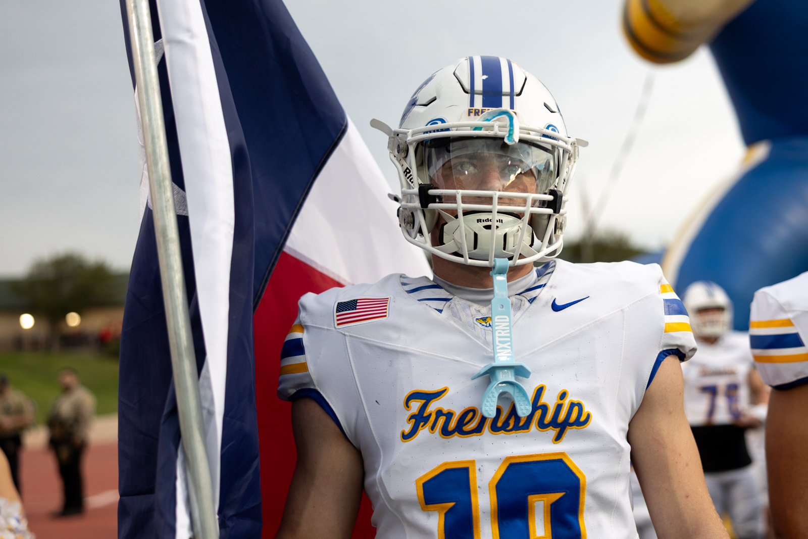 007A5628 | Press Pass Sports American football player in white jersey with blue and yellow trim stands with a flag draped behind him, helmet on and mouthguard visible.