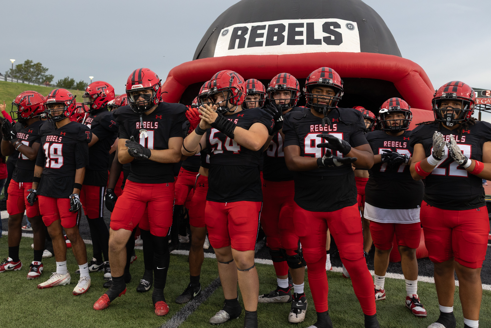 007A5643 | Press Pass Sports Football team in black jerseys and red pants standing under a red inflatable tunnel labeled REBELS before a game
