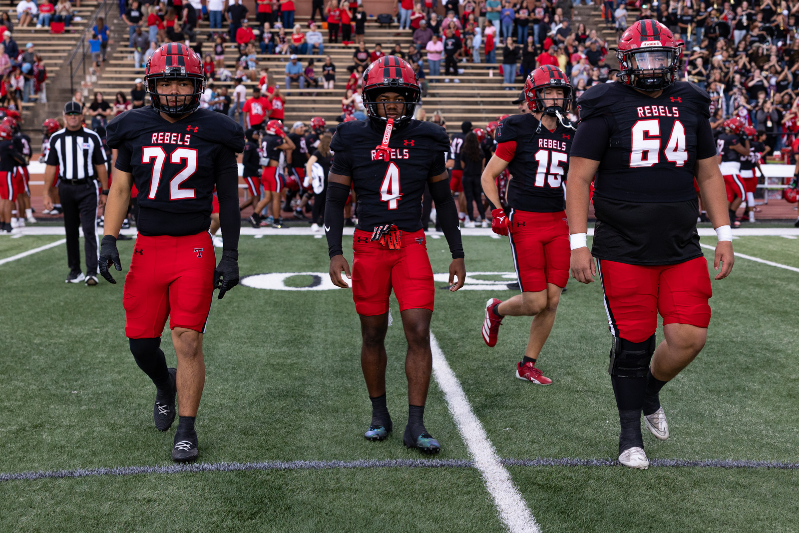 007A5657 | Press Pass Sports Rebels football players walk toward the camera on a green turf field, numbers 72, 4, 15, and 64, with fans in the stands behind them.