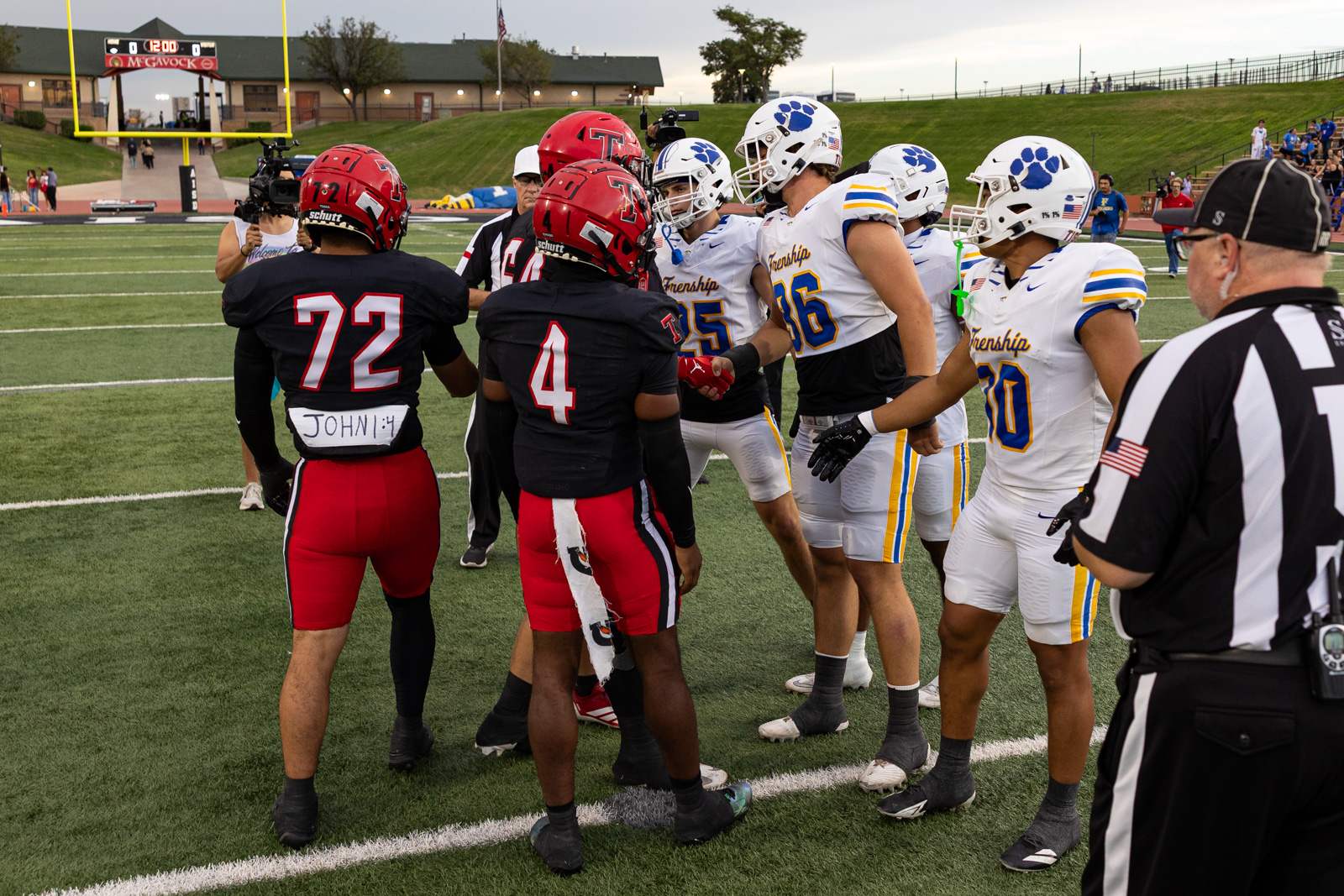 007A5671 | Press Pass Sports Two football teams greet each other at midfield before kickoff; red-and-black helmets contrast with white-and-blue uniforms, with a referee nearby in view.