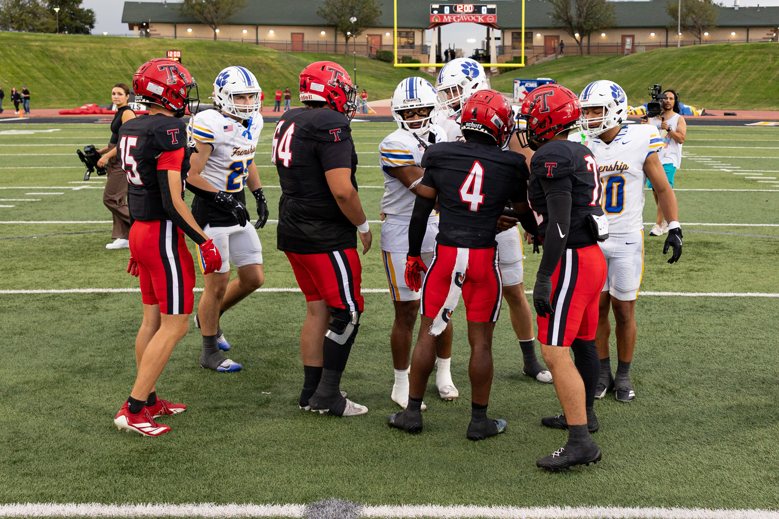 007A5690 | Press Pass Sports Football players from red-and-black and white-and-blue teams huddle at midfield on a turf field before the game, goalposts in background.