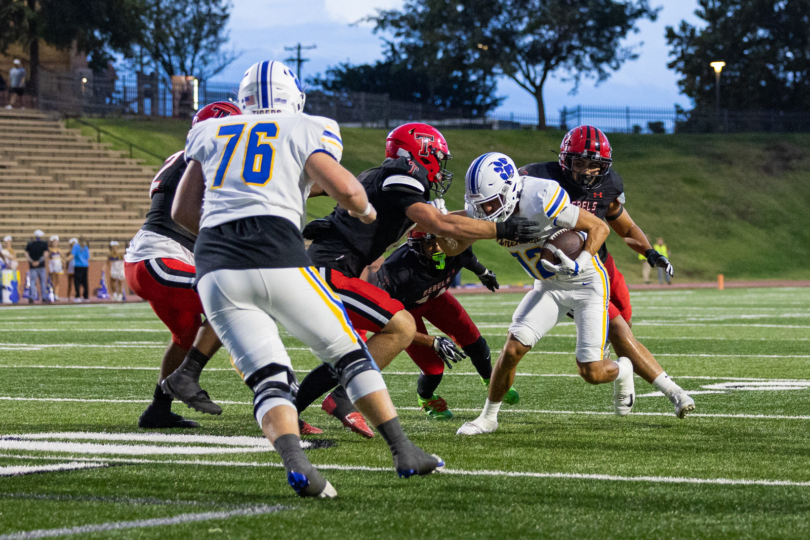011A5679 | Press Pass Sports Football play on a turf field: a ball carrier in white and blue attempts to run past a tackler from the opposing team in black and red.
