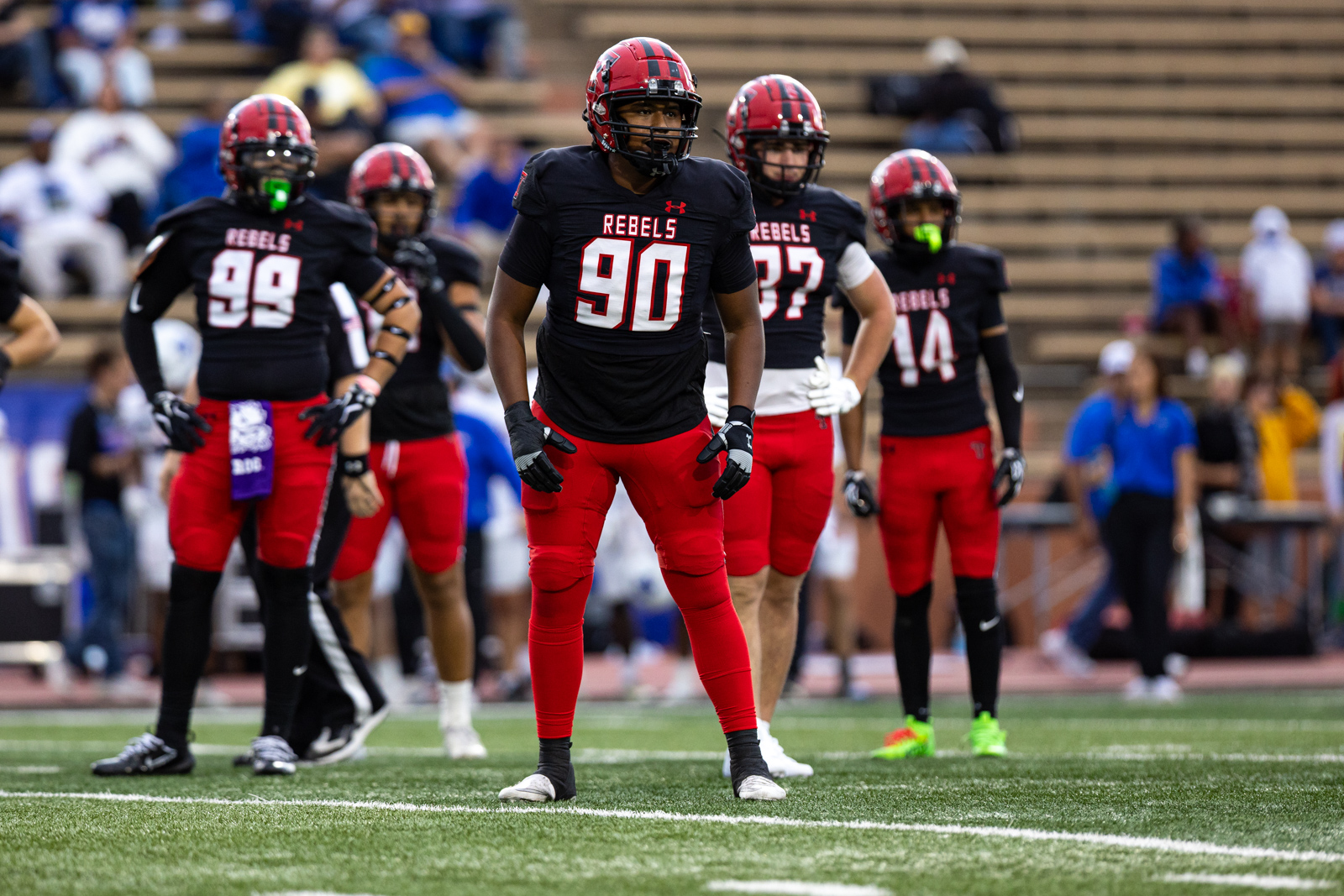 012A5278 | Press Pass Sports Center-stage football player wearing #90 in black jersey and red pants stands on the field with teammates in the background.