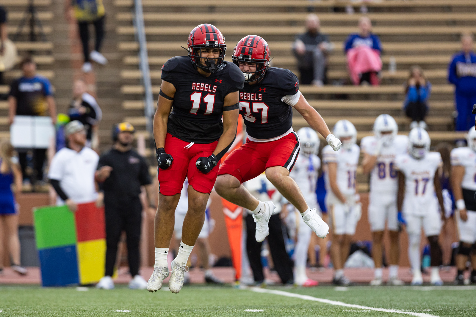 012A5324 | Press Pass Sports Two football players in black shirts with red numbers 11 and 37 leap into the air on a football field, cheering as teammates and spectators watch from the stands.