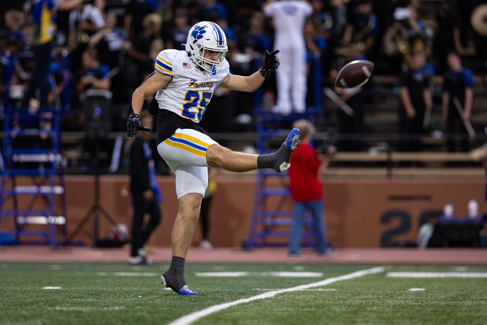 012A5395 | Press Pass Sports Football player in white and blue uniform kicking a ball mid-air on a turf field with a crowd in the background.