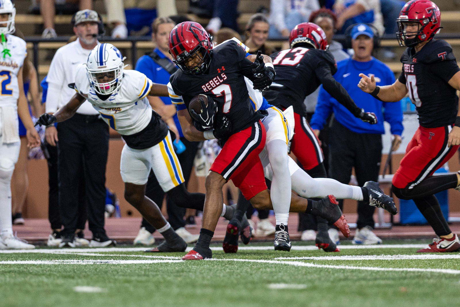 012A5674 | Press Pass Sports Football player in a black Rebels jersey (#7) carries the ball while being tackled by a white-uniform defender on a stadium field.
