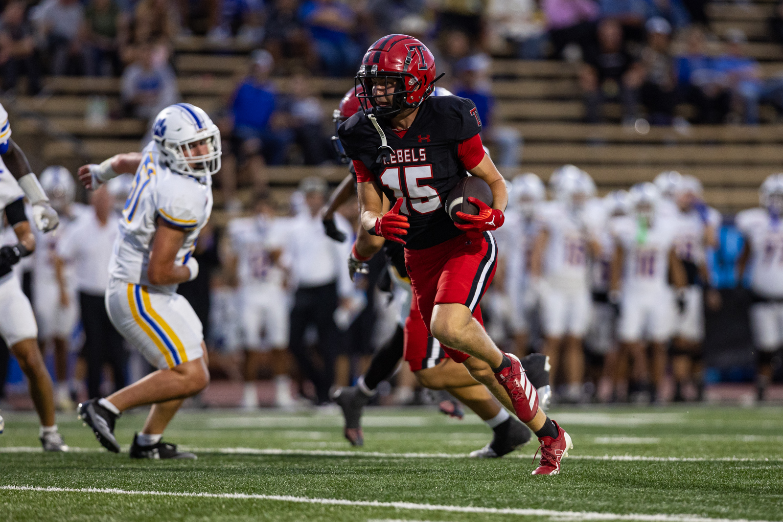 012A5751 | Press Pass Sports Football player in red-and-black Rebels uniform (#15) runs with the ball as a defender in white pursues him on the field.