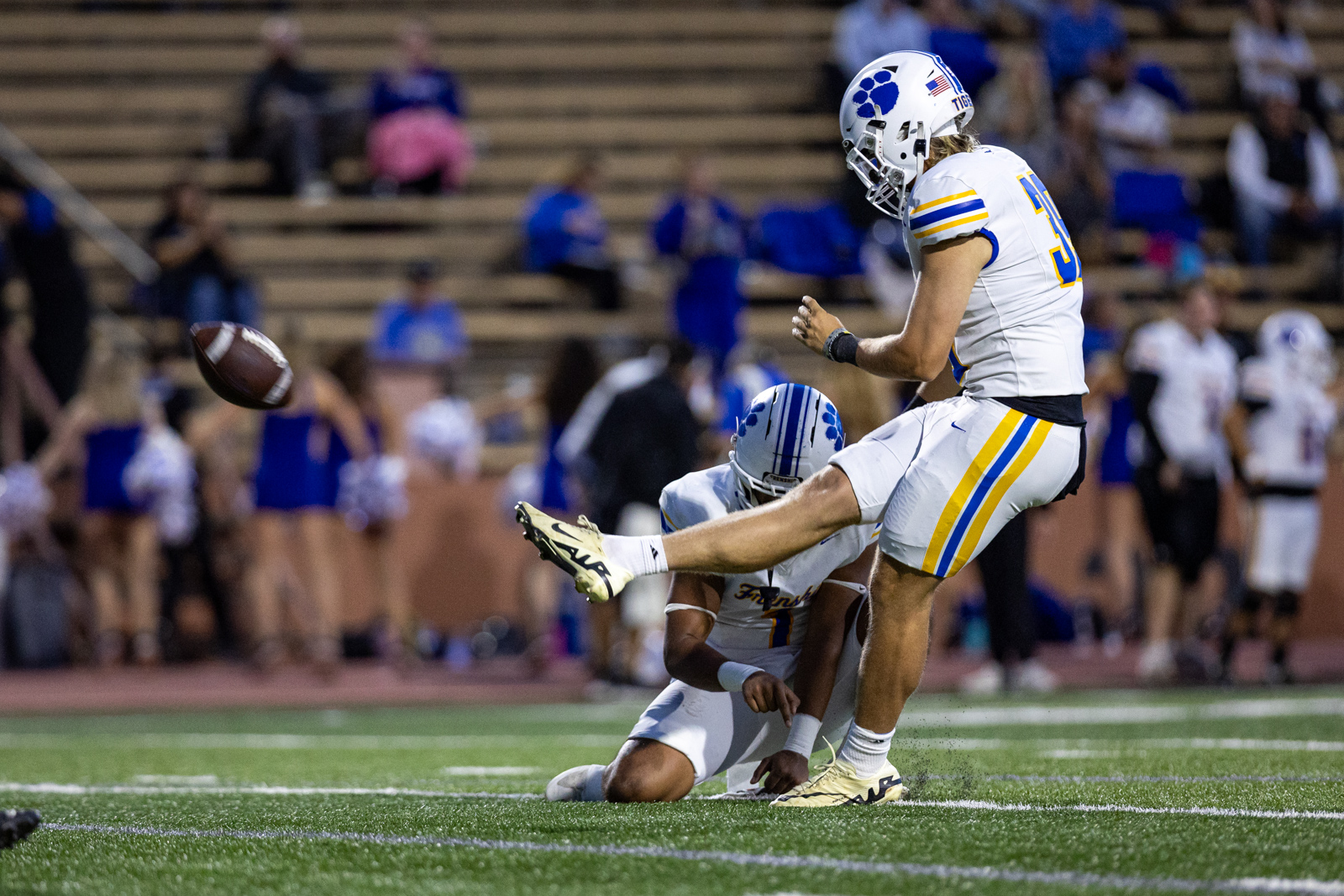 012A6005 | Press Pass Sports Football player in white and blue-yellow uniform kicks a football while a teammate in white lies on the turf nearby; stadium spectators in the background.