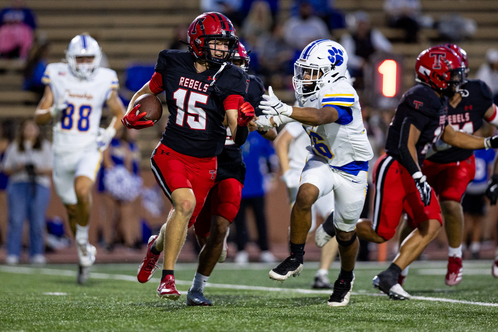 012A6054 | Press Pass Sports Rebels running back wearing #15 in black and red carries the ball while a defender in white and blue reaches in a crowded football field.