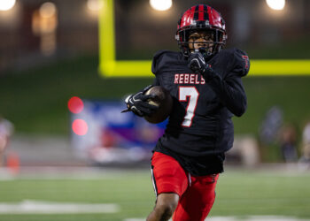 012A6182 | Press Pass Sports Football player wearing a black Rebels jersey with number 7 dribbles the ball while sprinting on a green field at night stadium.