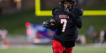 Football player wearing a black Rebels jersey with number 7 dribbles the ball while sprinting on a green field at night stadium.