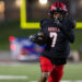 012A6182 | Press Pass Sports Football player wearing a black Rebels jersey with number 7 dribbles the ball while sprinting on a green field at night stadium.