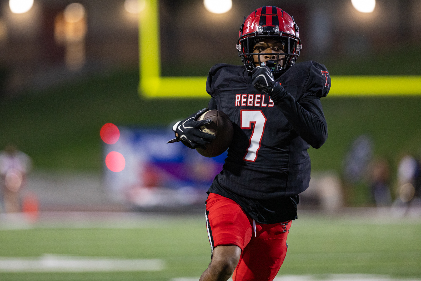 012A6182 | Press Pass Sports Football player wearing a black Rebels jersey with number 7 dribbles the ball while sprinting on a green field at night stadium.