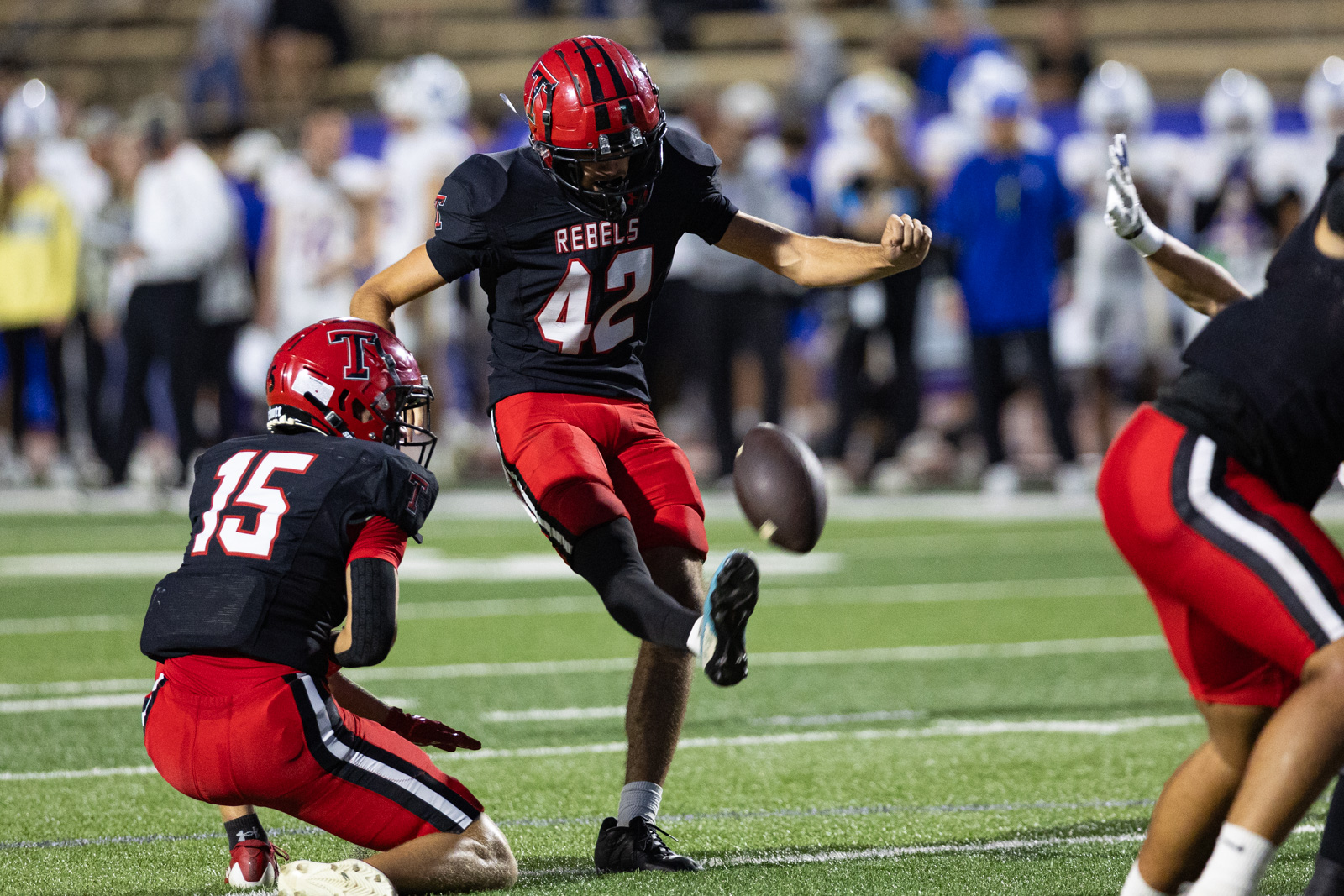 012A6281 | Press Pass Sports Football players in black and red uniforms on a green turf field; #42 jumps as the ball rolls loose nearby while teammate #15 prepares to block or pursue.