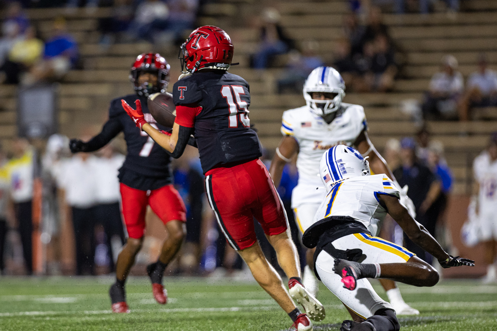 012A6444 | Press Pass Sports Football players in a night game: a red-helmeted player wearing number 15 in a black uniform reaches for the ball while a defender in white and blue pursues nearby on a football field behind him.