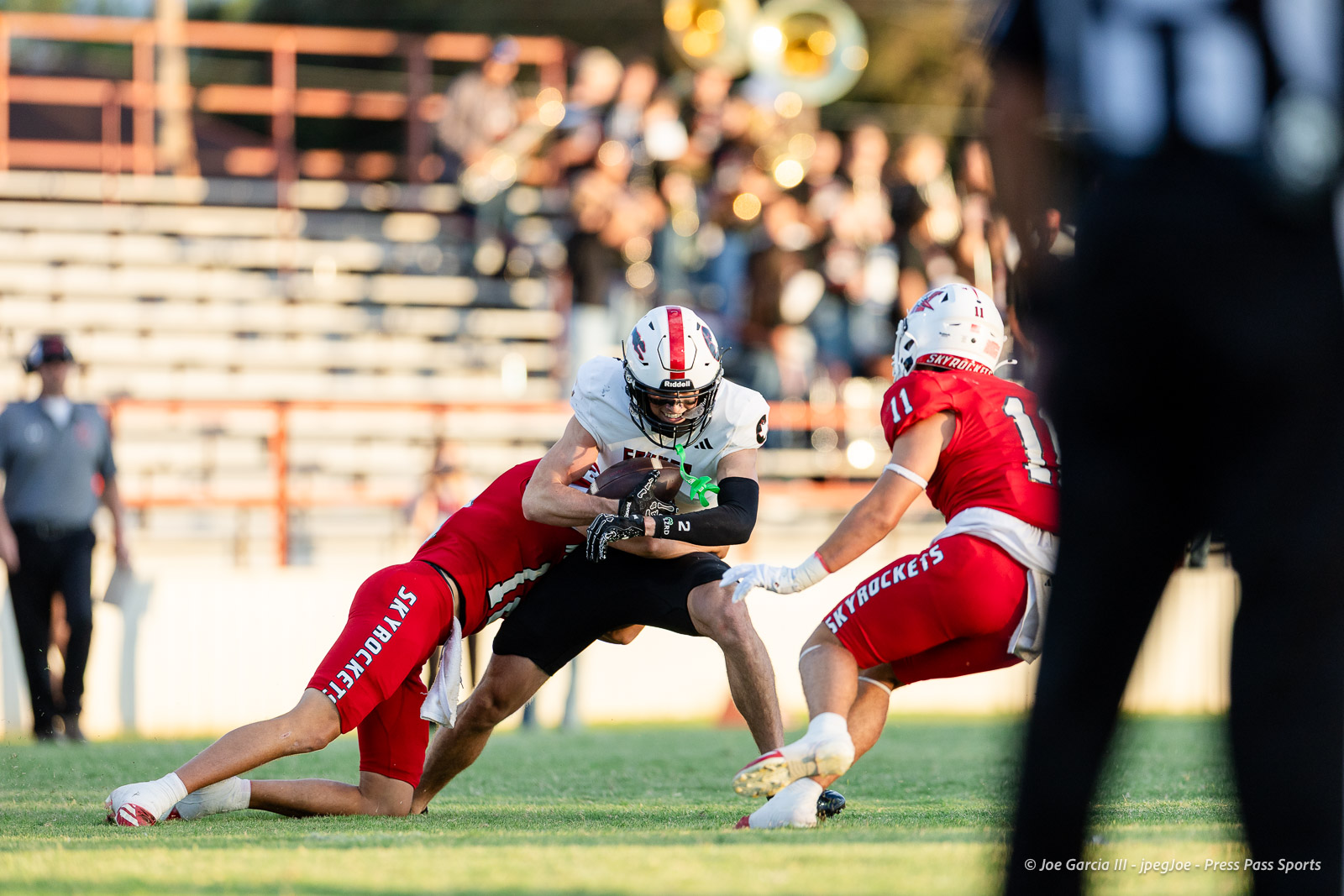 gruver-vs-wellinton-2026-jpegJoe-pps-2 | Press Pass Sports White-jersey football player cradles the ball while being tackled by a red-clad defender on a sunlit field.