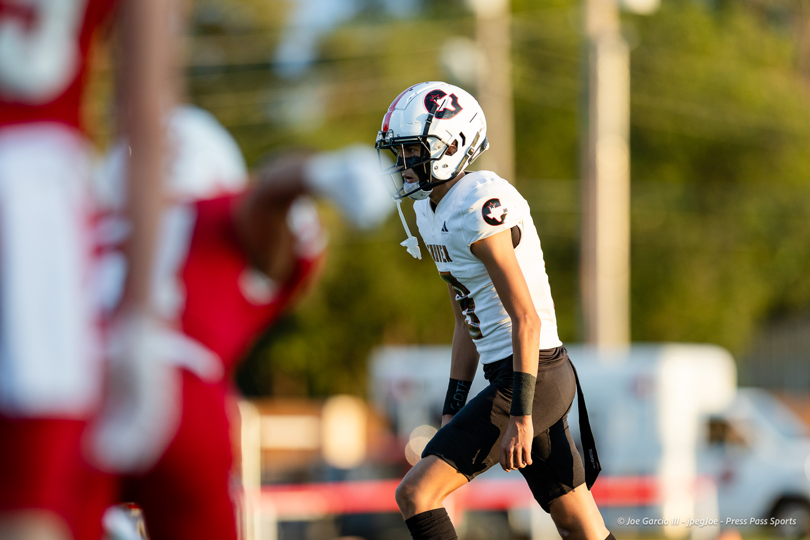 gruver-vs-wellinton-2026-jpegJoe-pps-5 | Press Pass Sports Football player in a white uniform and helmet on the field during a game, with red-clad players blurred in the foreground