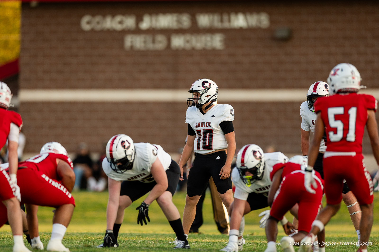 gruver-vs-wellinton-2026-jpegJoe-pps-8 | Press Pass Sports High school football game at the line of scrimmage, players in white and red jerseys facing off on a sunny field with a brick fieldhouse in the background.