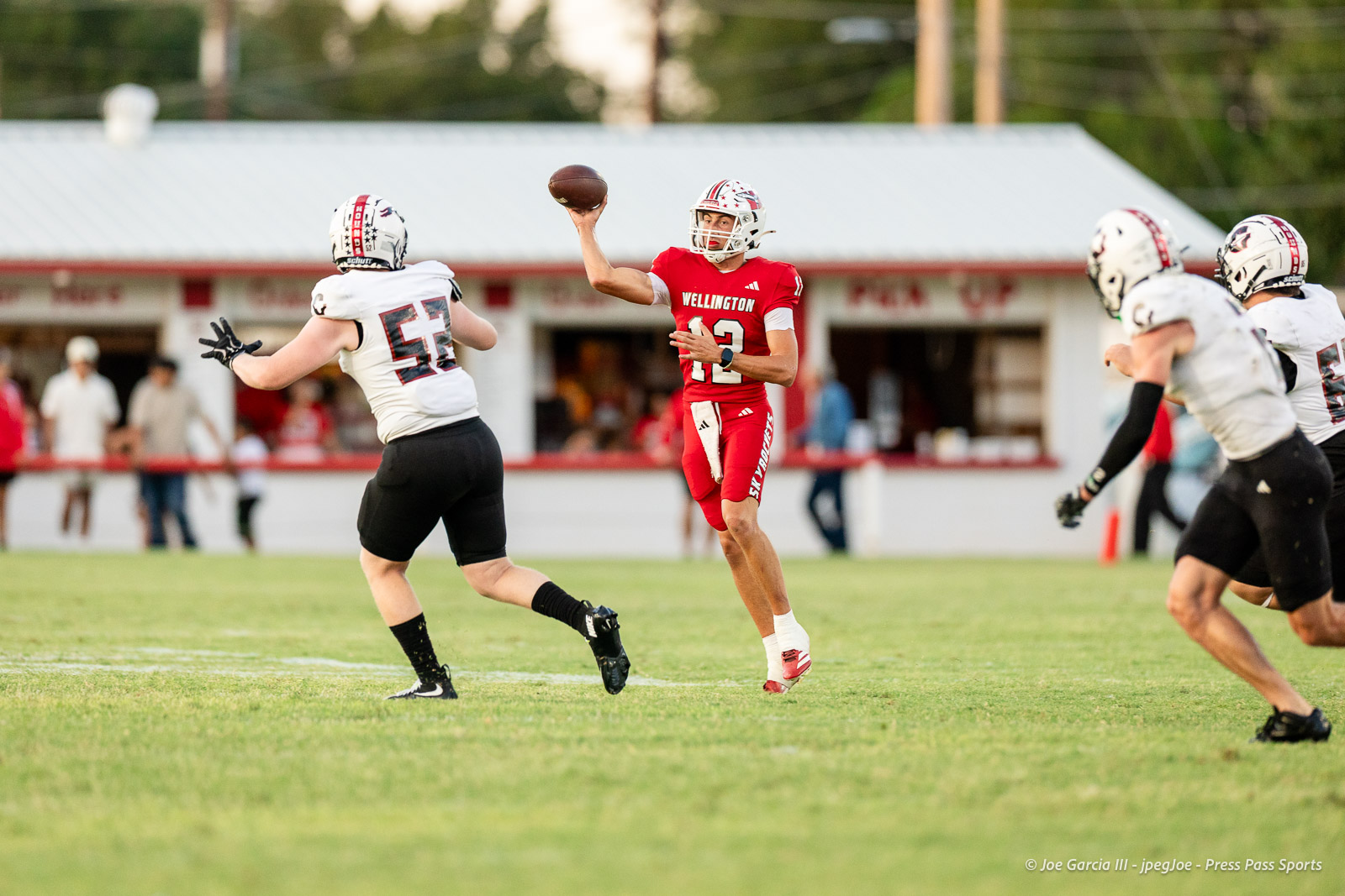 gruver-vs-wellinton-2026-jpegJoe-pps-9 | Press Pass Sports Quarterback in red Wellington uniform throws a pass while defenders approach on a grassy football field.