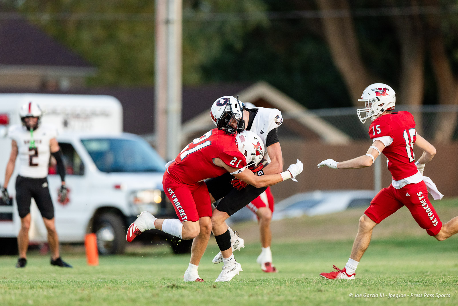 gruver-vs-wellinton-2026-jpegJoe-pps-10 | Press Pass Sports Two players in red uniforms tackle a white-uniformed player during a football game on a grassy field; a teammate approaches from the right.