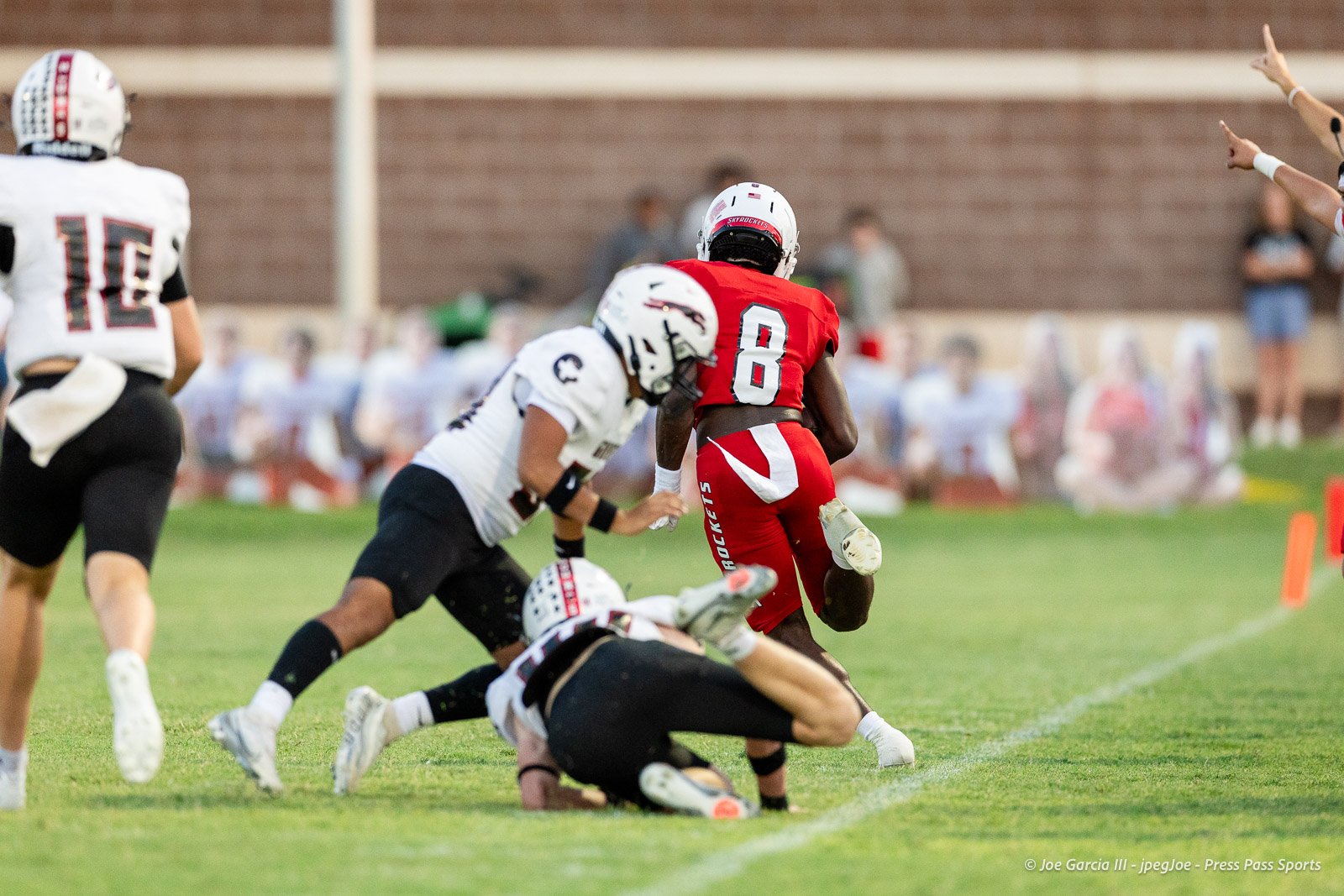 gruver-vs-wellinton-2026-jpegJoe-pps-11 | Press Pass Sports Red-jersey football running back wearing #8 carries the ball as white-jersey defenders close in on a grassy field with a blurred crowd in the background.