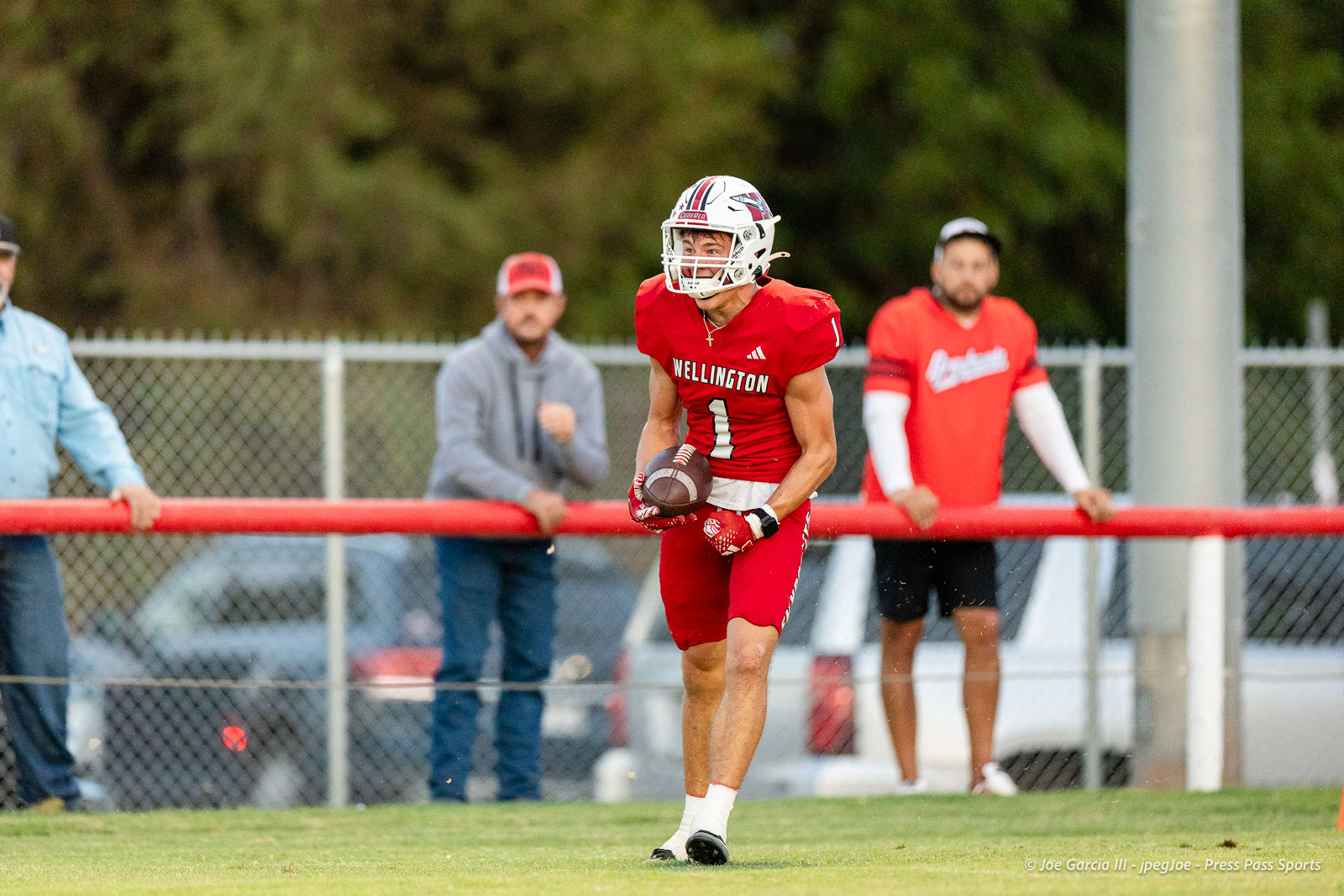 gruver-vs-wellinton-2026-jpegJoe-pps-12 | Press Pass Sports Football player in red Wellington uniform running with ball during a play, spectators behind a red barrier in a fenced field.