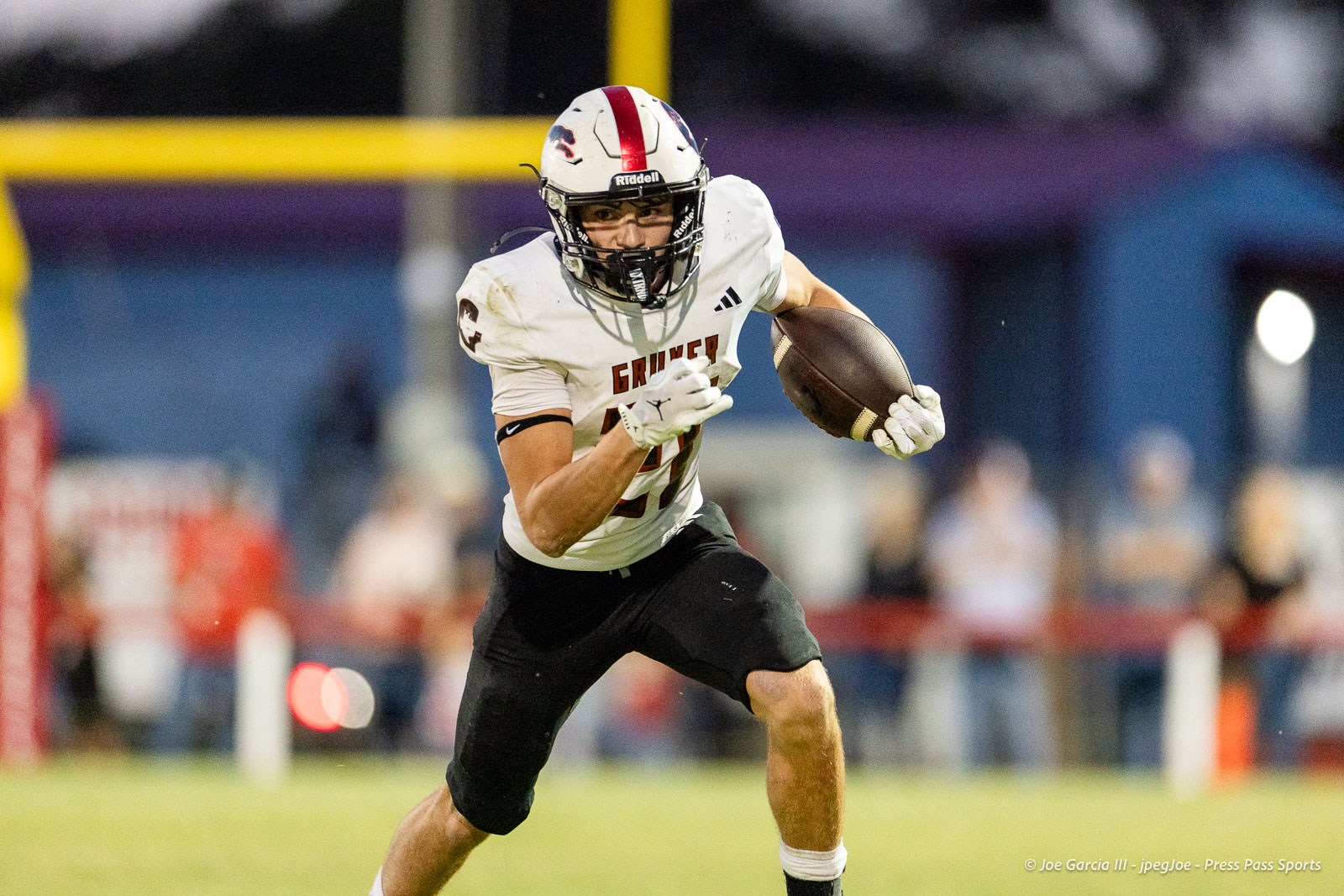 gruver-vs-wellinton-2026-jpegJoe-pps-13 | Press Pass Sports Football player in a white jersey runs with a football tucked under his arm on a grassy field during a game; blurred spectators in the background.