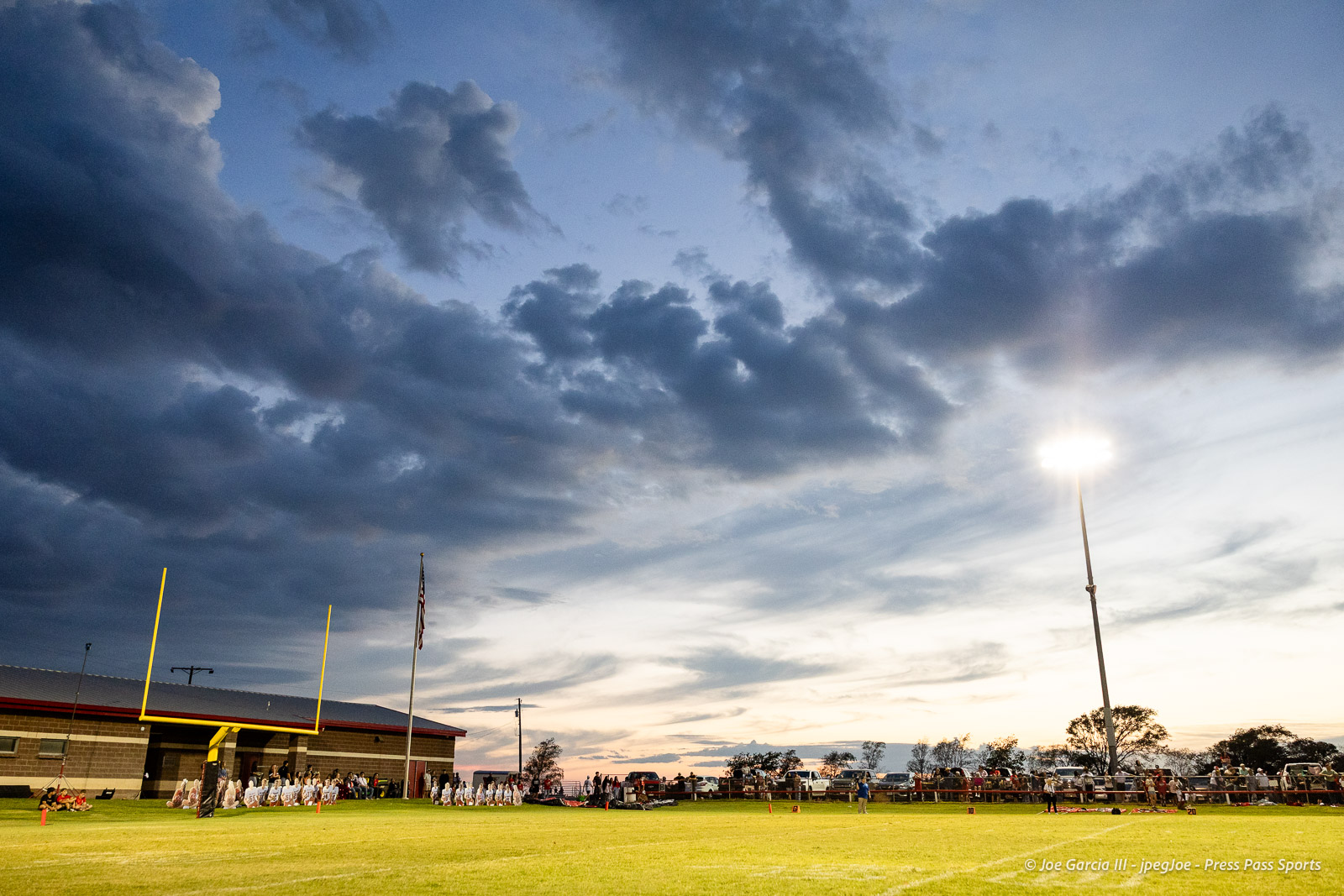 gruver-vs-wellinton-2026-jpegJoe-pps-14 | Press Pass Sports Evening football field with a bright floodlight, yellow goalposts, and spectators along the sideline under a cloudy sky.