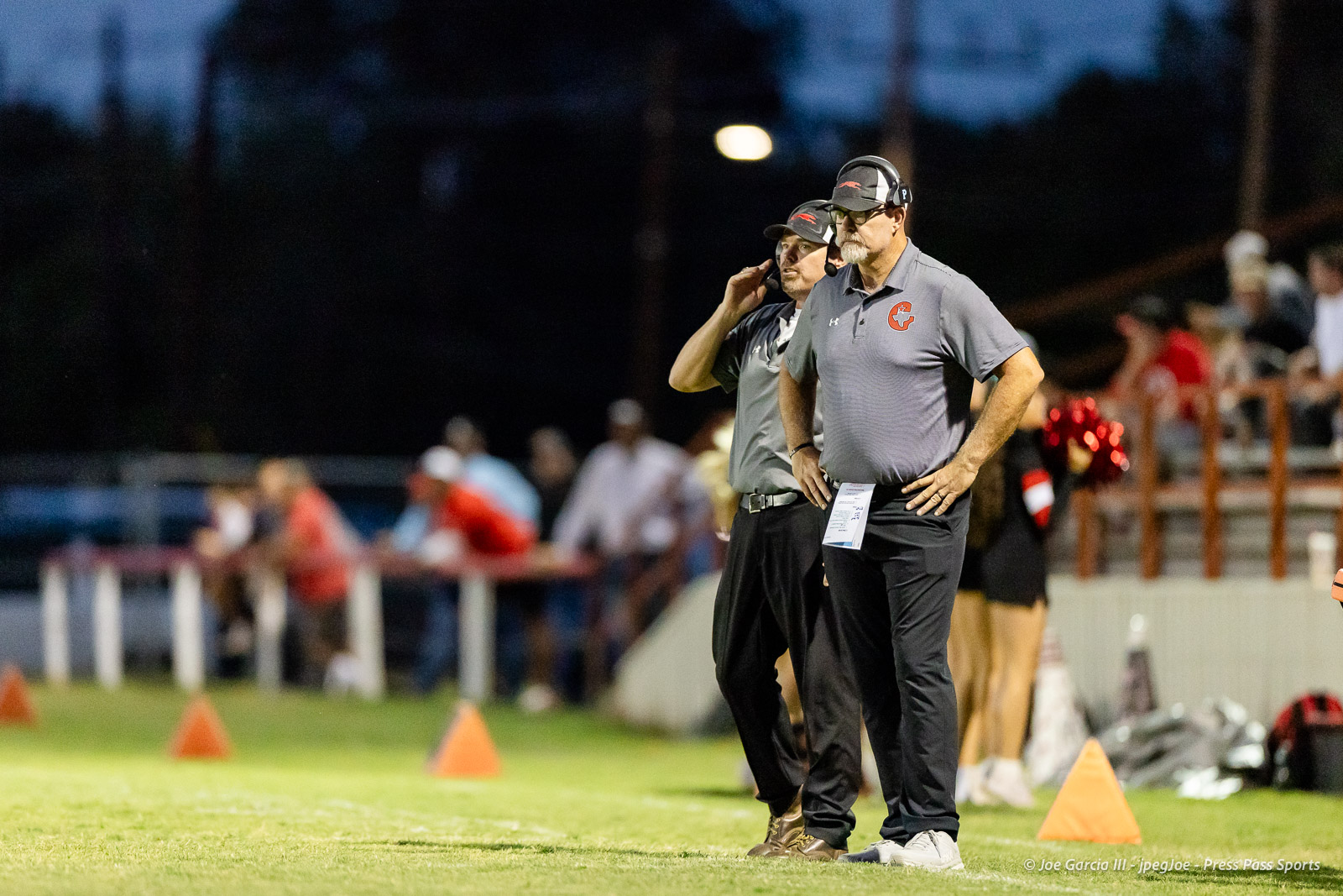 gruver-vs-wellinton-2026-jpegJoe-pps-16 | Press Pass Sports Two coaches in matching gray polo shirts and headsets stand on the sideline during a nighttime football game, hands on hips and focused on the field.