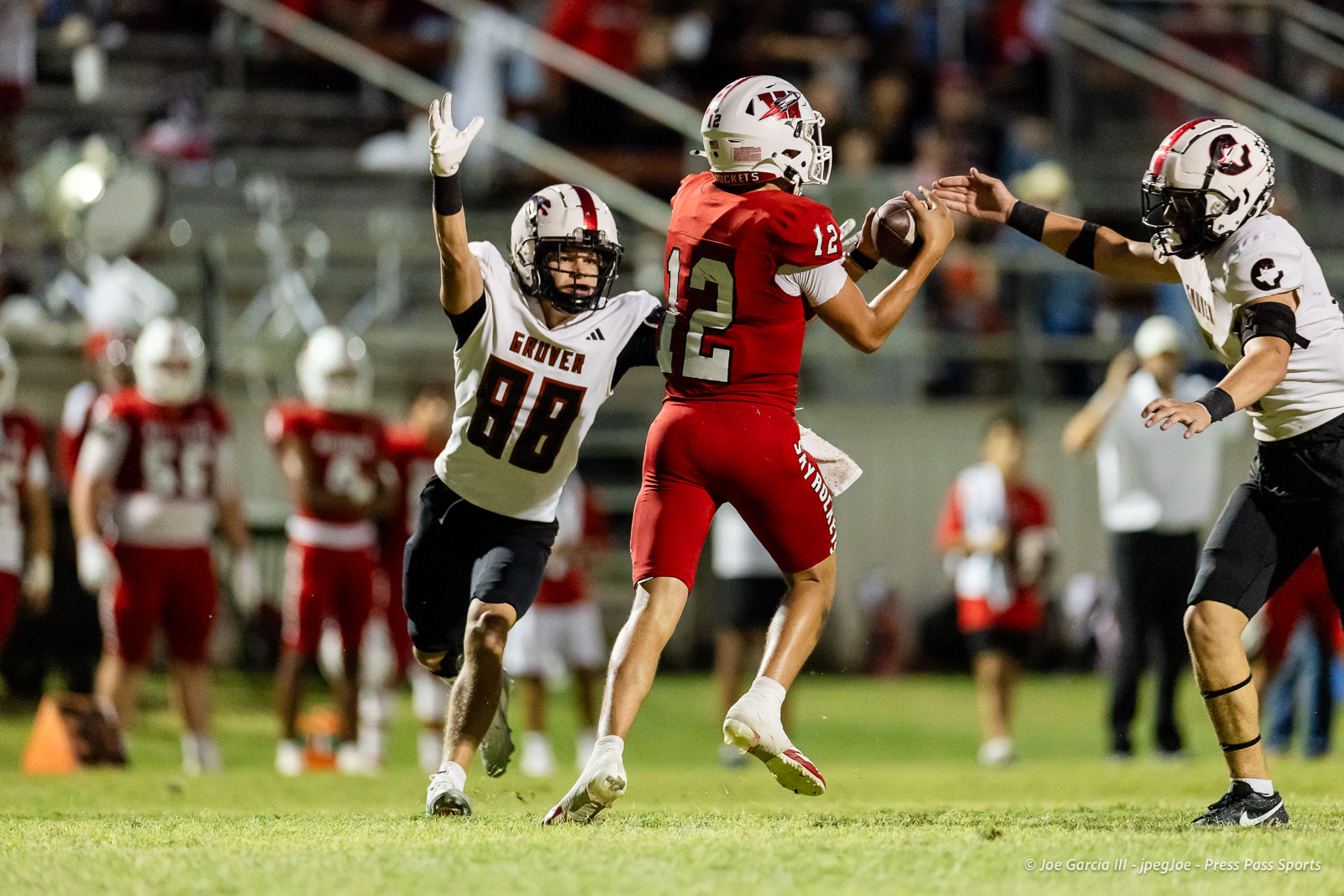 gruver-vs-wellinton-2026-jpegJoe-pps-17 | Press Pass Sports Football player in a red jersey (No. 12) catches a pass while a white-jersey defender (No. 88) reaches to disrupt him on a grassy field under stadium lights.