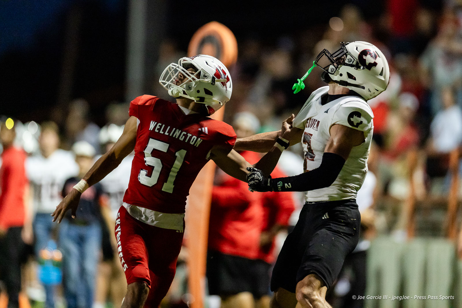 gruver-vs-wellinton-2026-jpegJoe-pps-18 | Press Pass Sports Two football players collide during a game: a Wellington defender in red #51 blocks a white‑jersey opponent as the crowd looks on, night lights in background.