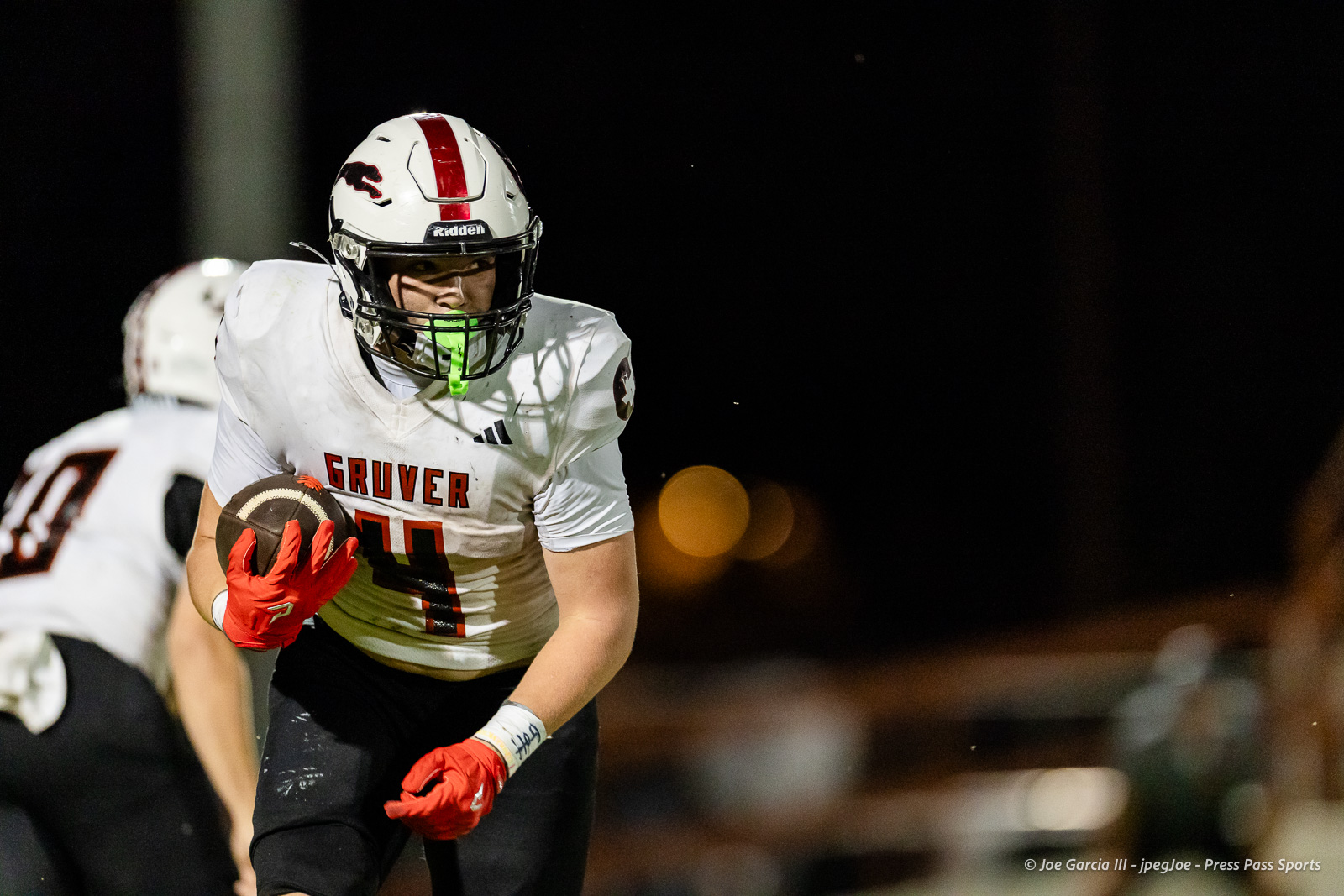 gruver-vs-wellinton-2026-jpegJoe-pps-19 | Press Pass Sports High school football player in a white Gruver jersey (#11) runs with the ball at night, wearing red gloves and a neon-green mouthguard in a dark stadium.