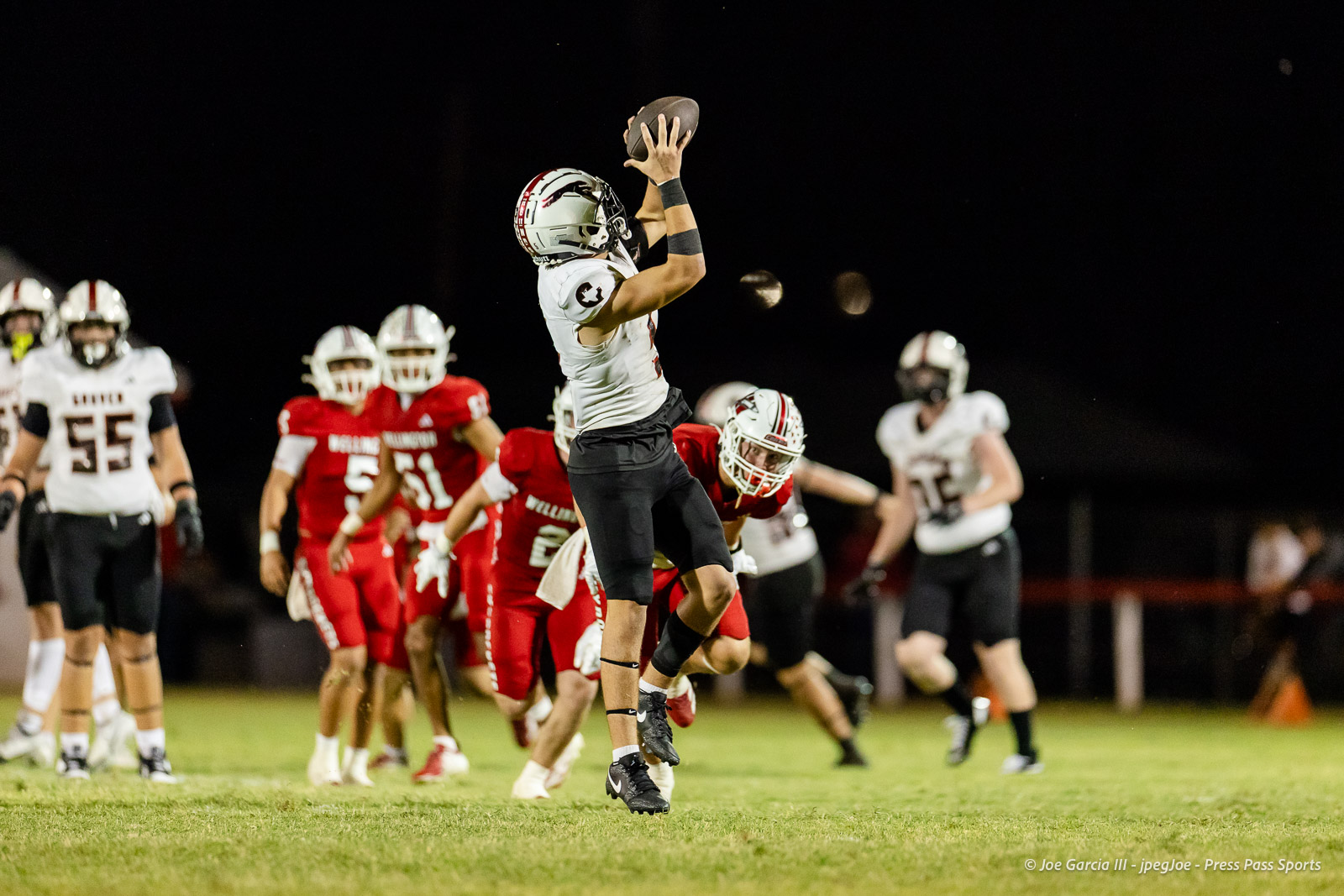 gruver-vs-wellinton-2026-jpegJoe-pps-22 | Press Pass Sports A football player in a white jersey leaps to catch a pass while defenders in red close in at night on a grassy field.