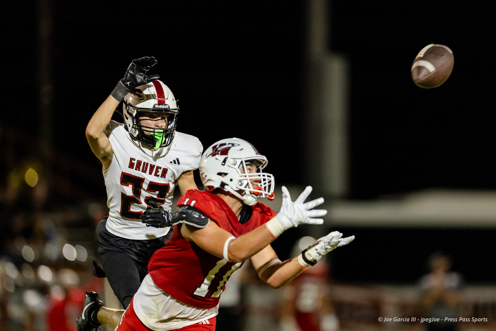 gruver-vs-wellinton-2026-jpegJoe-pps-24 | Press Pass Sports Two high school football players in a night game reach for the football; white Gruver jersey #23 vs a red opponent.