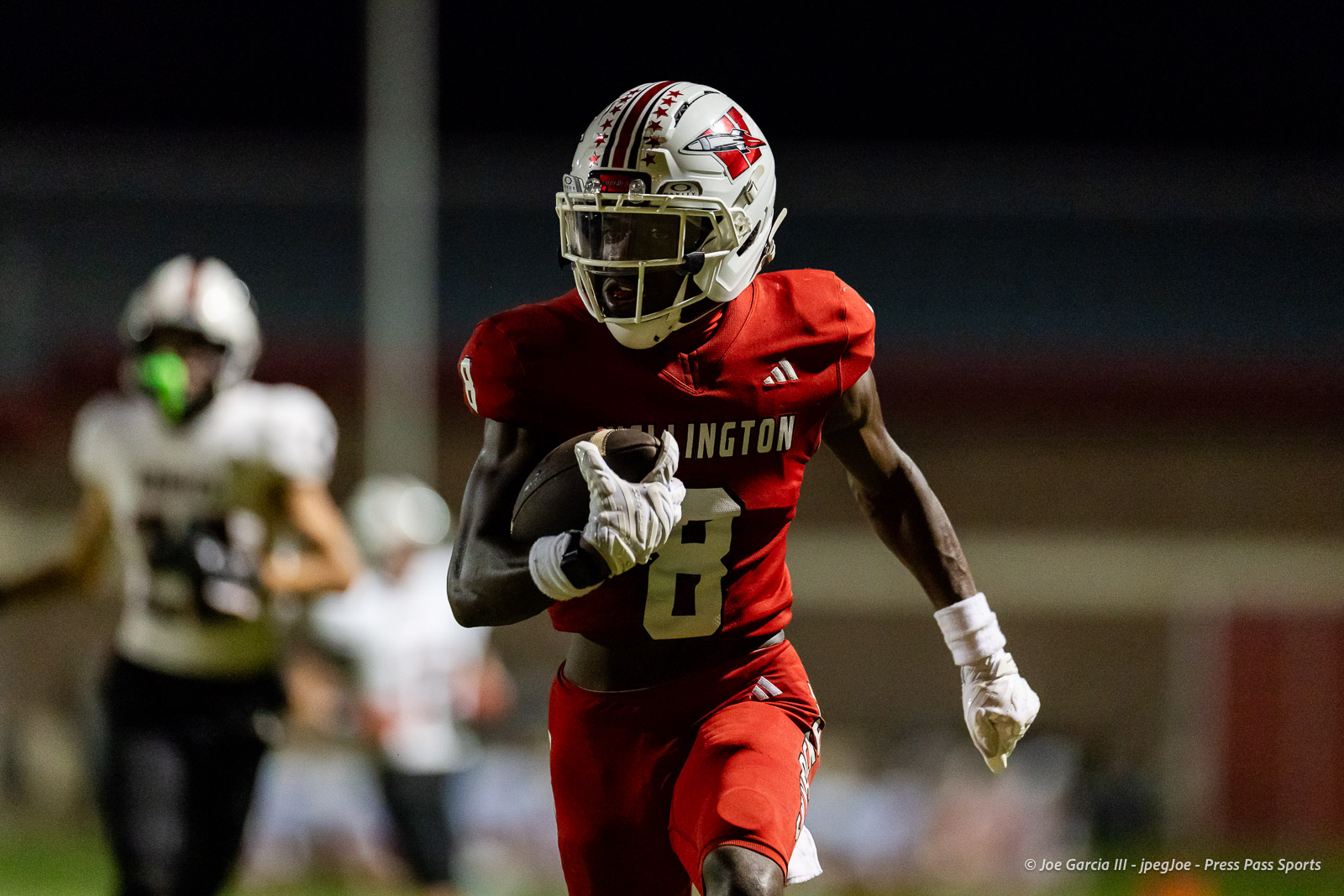 gruver-vs-wellinton-2026-jpegJoe-pps-26 | Press Pass Sports Football player in a red uniform (#8) runs with the ball during a night game, helmet and white gloves visible.