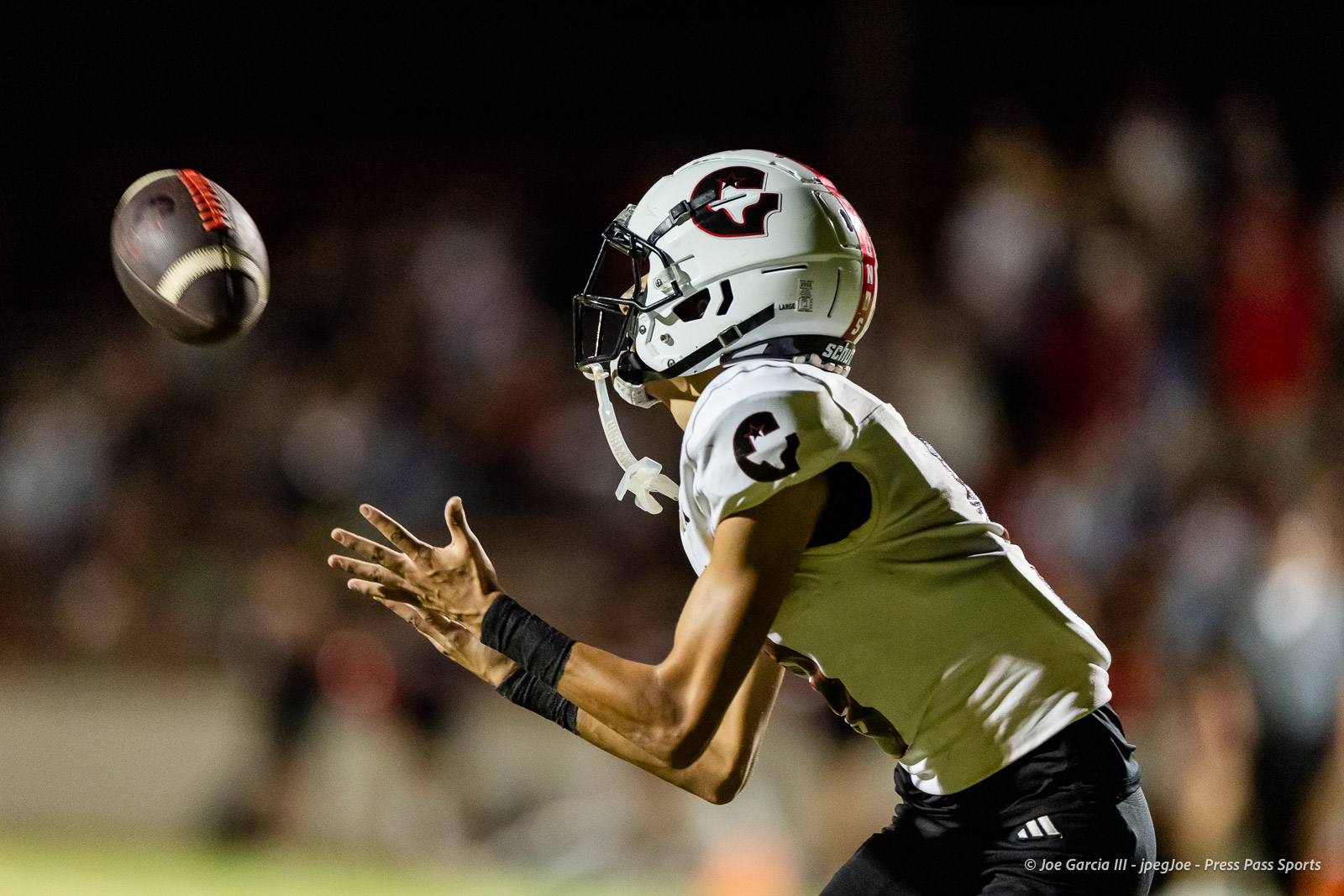 gruver-vs-wellinton-2026-jpegJoe-pps-27 | Press Pass Sports American football player in a white jersey and helmet reaches for a spiraling ball during a night game with a blurred crowd in the background.