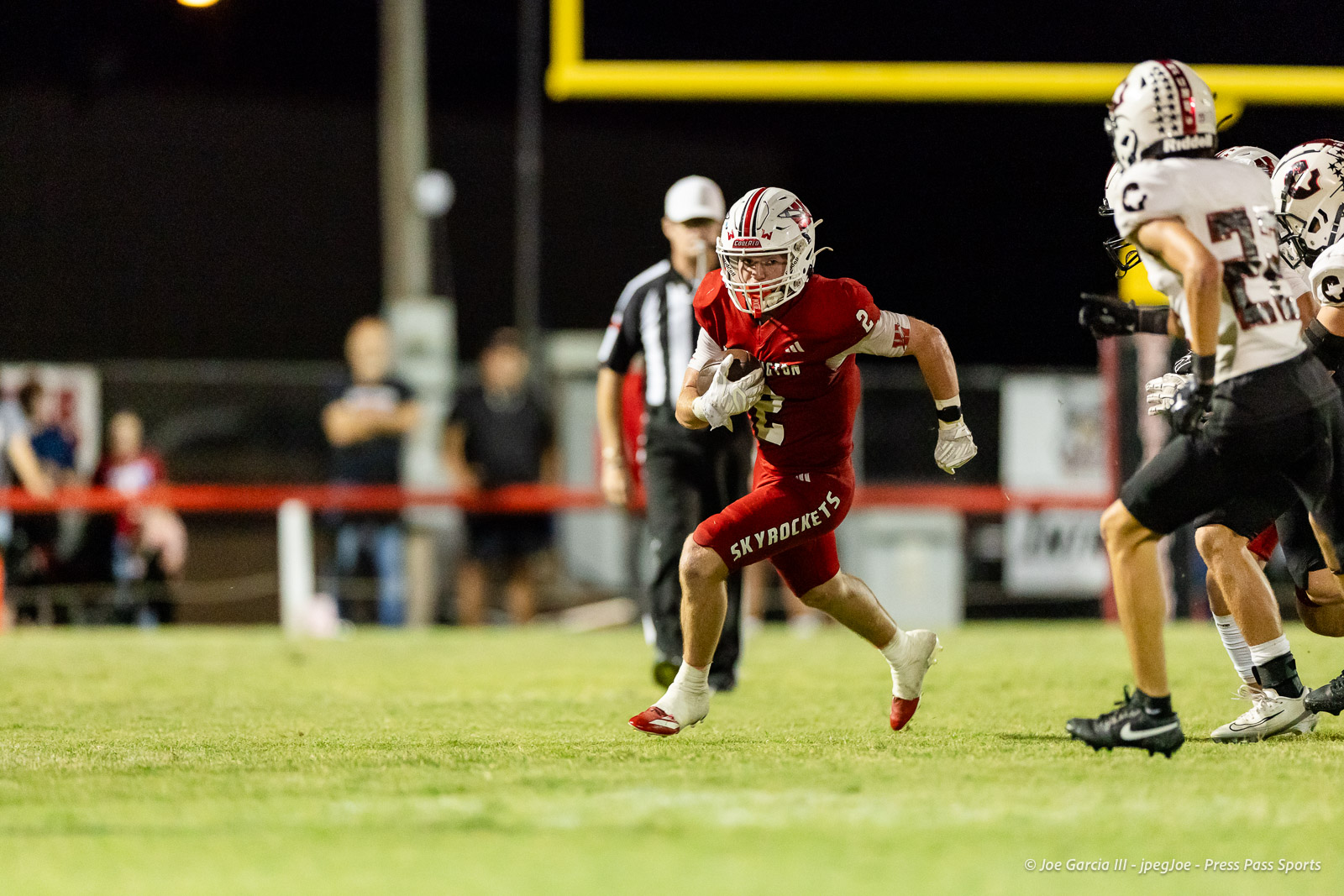 gruver-vs-wellinton-2026-jpegJoe-pps-30 | Press Pass Sports Football player in red jersey carrying the ball, sprinting toward the end zone during a night game; others in white uniforms pursue him.