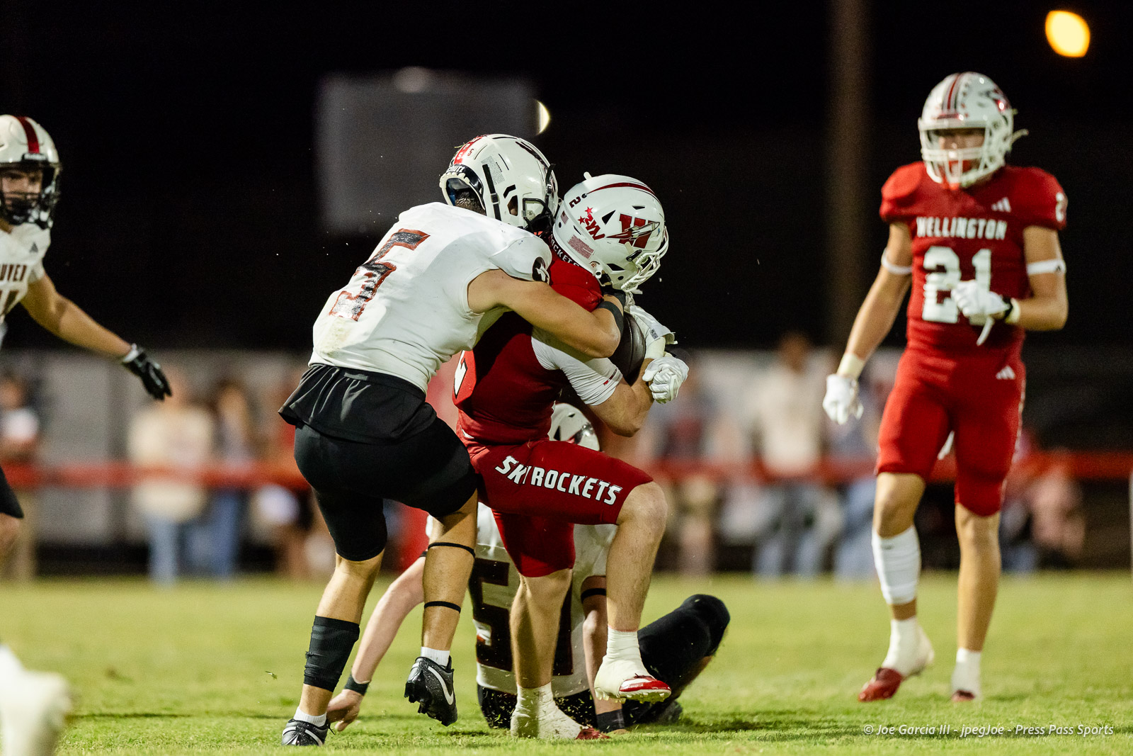 gruver-vs-wellinton-2026-jpegJoe-pps-31 | Press Pass Sports Rugby-like football play at night: white-jersey players tackle a red-jersey ball carrier on a grassy field.